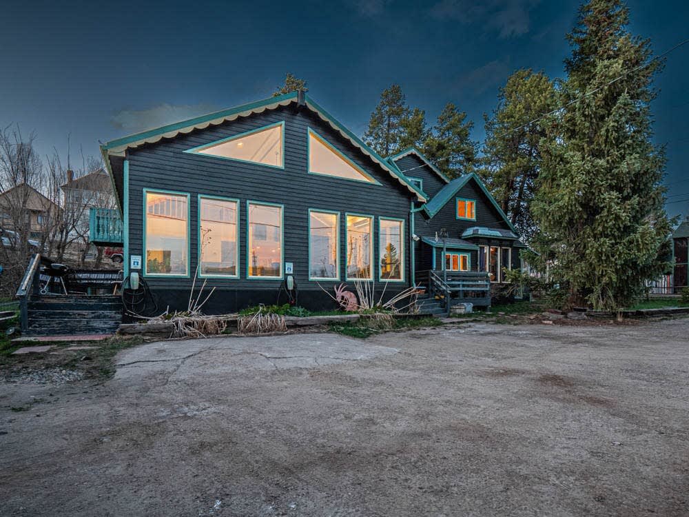 A large, dark-colored house features multiple expansive windows that illuminate the exterior. The surrounding area is marked by gravel and a few plants, with two Tesla EV charging stations visible on the side of the structure. Trees provide a natural backdrop against the evening sky.