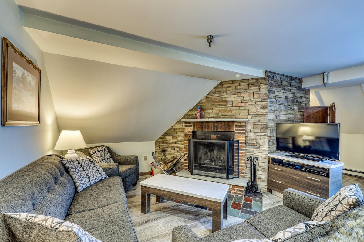 A cozy living area is featured, showcasing a sectional sofa and two armchairs arranged around a coffee table. A stone fireplace serves as a focal point against the wall, with a television placed on a media console adjacent to it. Soft lighting is provided by a lamp in the corner.