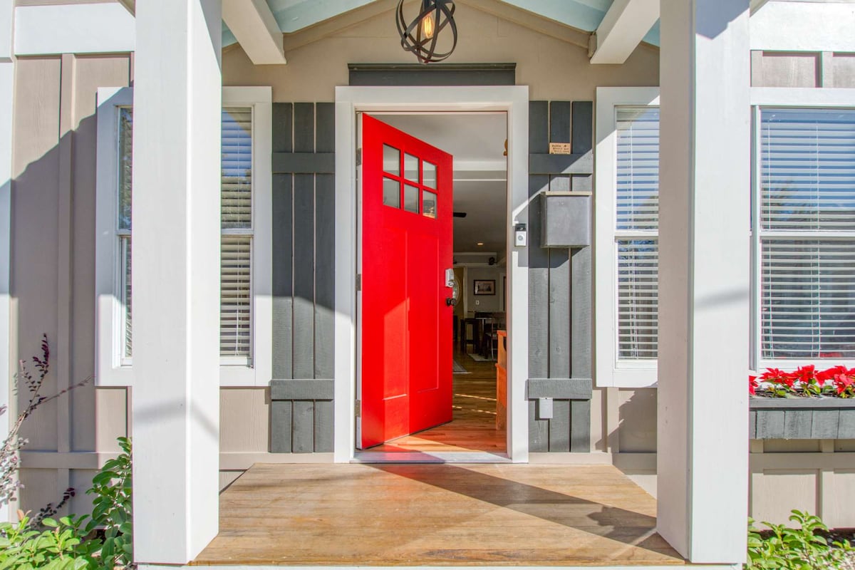A welcoming entrance features a bright red door framed by white columns. Natural light floods the entryway, highlighting the hardwood floor. Greenery flanks the entrance, and windows on either side allow for ample brightness inside.