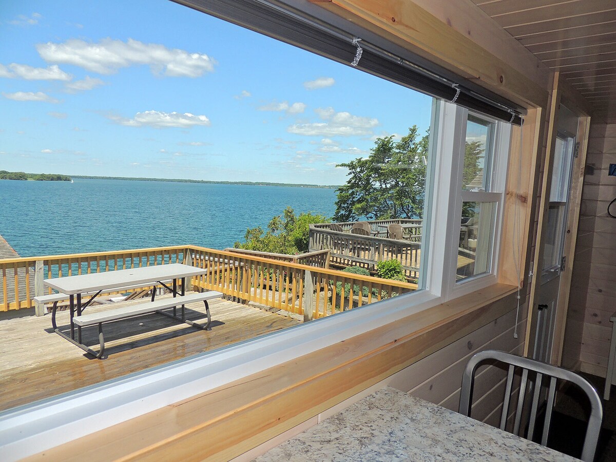 A large scenic window invites natural light into the cottage, framing a view of the St. Lawrence River. Outside, a picnic table is positioned on a spacious deck surrounded by greenery, enhancing the serene atmosphere of the waterfront setting.