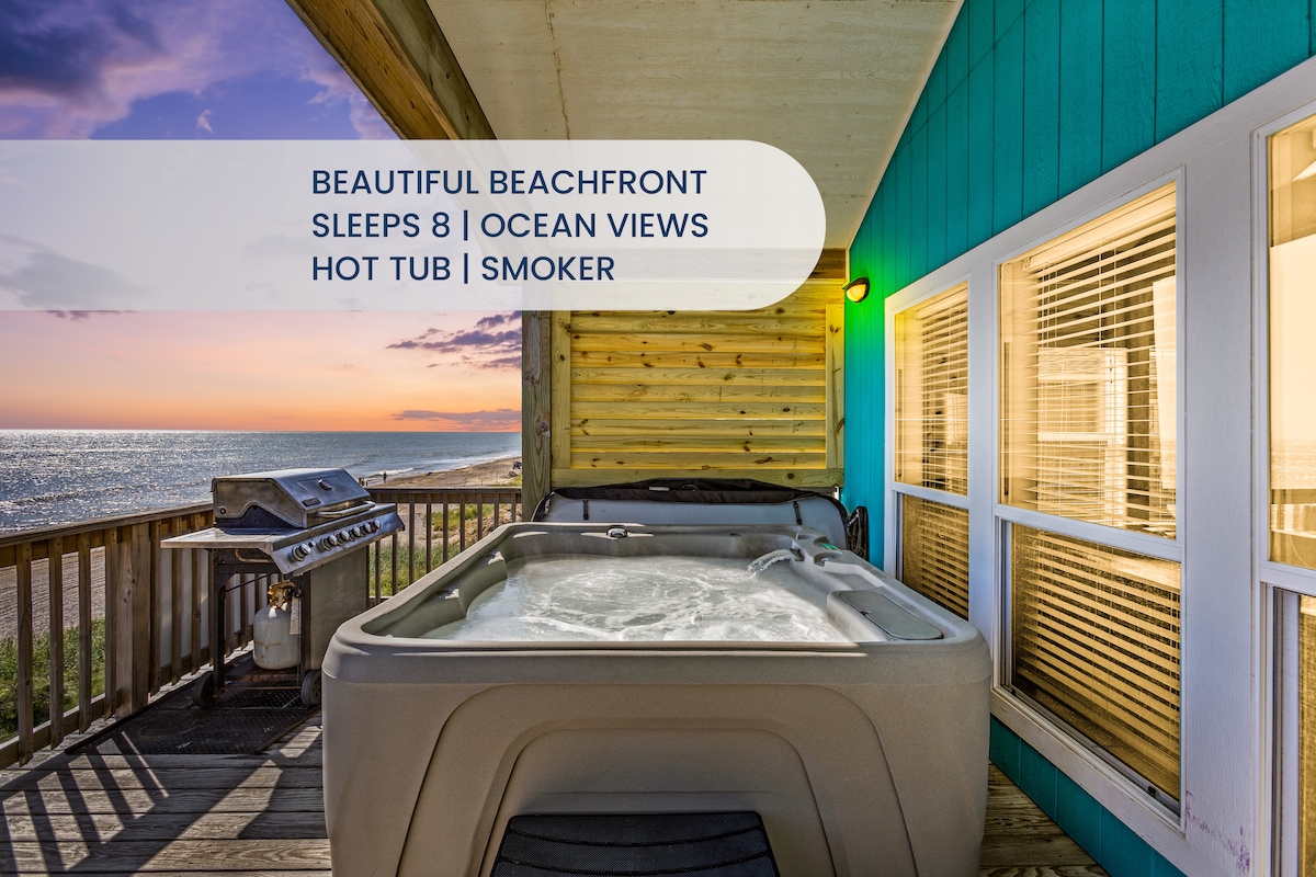 A hot tub is positioned on the spacious deck, surrounded by wooden beams and colorful siding. The ocean can be viewed in the distance, while a grill is available nearby for outdoor cooking. Light from the evening sky casts a warm glow over the area.