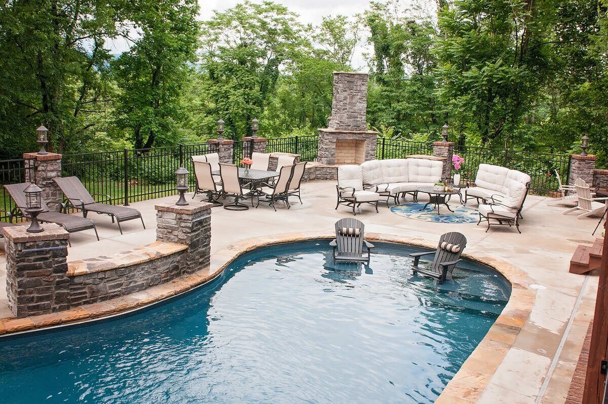 The pool area features a crystal-clear swimming pool surrounded by a natural stone patio. Comfortable lounge chairs and deep seating are arranged around the space, while a stone fireplace adds a rustic touch. Lush greenery frames the area, enhancing the overall tranquility.