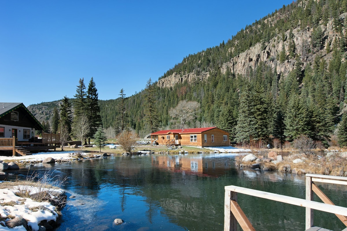 A tranquil scene depicts the Sequoia Cabin alongside a clear, reflective trout pond. Lush greenery surrounds the area, with tall trees rising in the background. Snow remains in patches, hinting at a crisp mountain atmosphere under a bright blue sky.