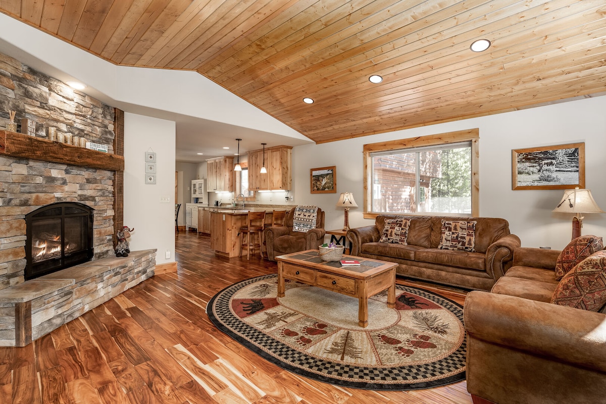 A spacious living area features a stone fireplace at the center, surrounded by two couches and a coffee table. Natural wood accents the ceiling, while a patterned area rug adds warmth. A window provides natural light, leading to a view of the kitchen beyond.