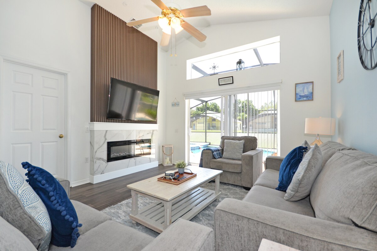 A spacious living area features a large flat screen television mounted above a modern fireplace. Comfortable gray sofas are arranged around a light coffee table. Natural light floods in through large windows, with views of the sunny pool area visible in the background.