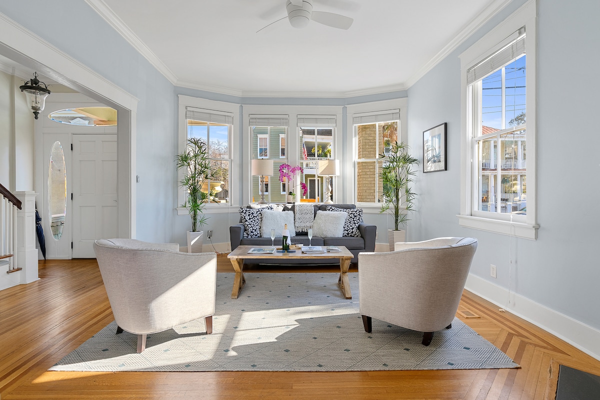 The living area showcases a bright, airy space adorned with large windows allowing natural light to flood in. A cozy seating arrangement includes a sofa and two armchairs, with a patterned area rug beneath a central coffee table. Potted plants add a touch of greenery.
