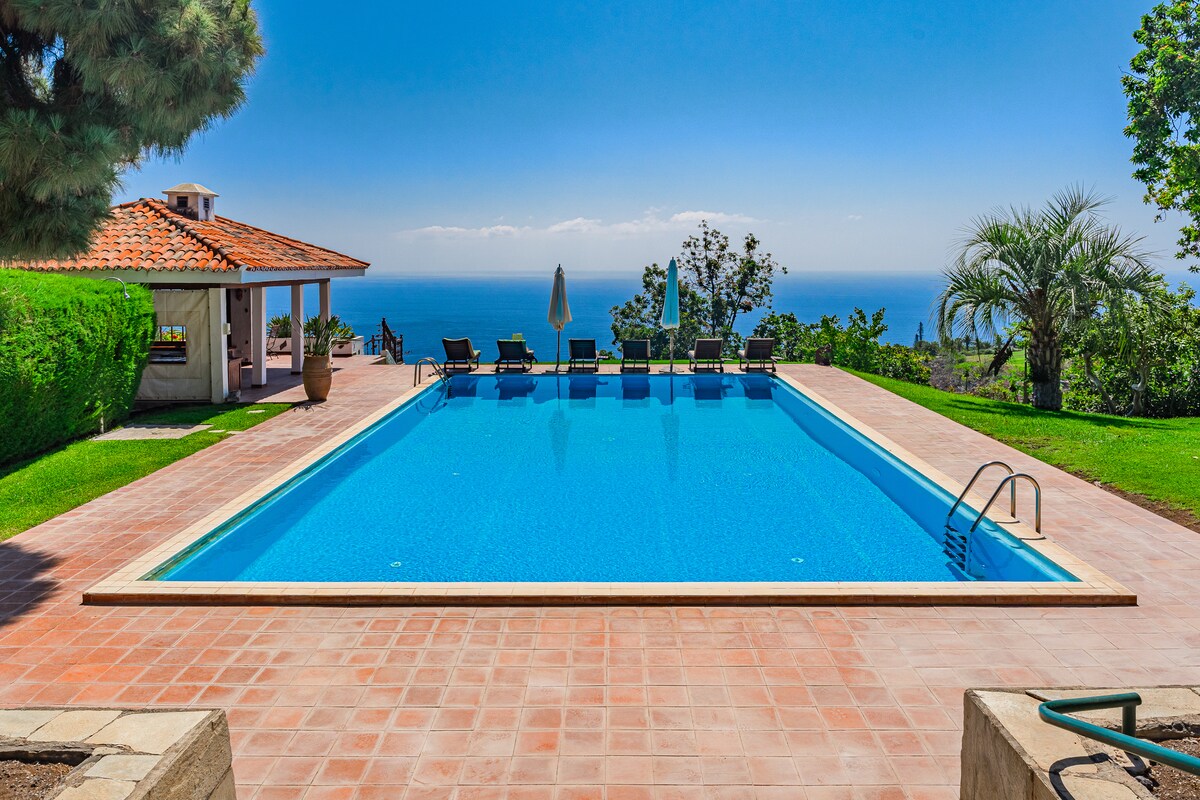 A spacious swimming pool is framed by a well-manicured lawn and trees, offering views of the ocean. Several sun loungers line the poolside, providing relaxation options. A shaded area with a parasol and the villa's structure can be seen in the background.