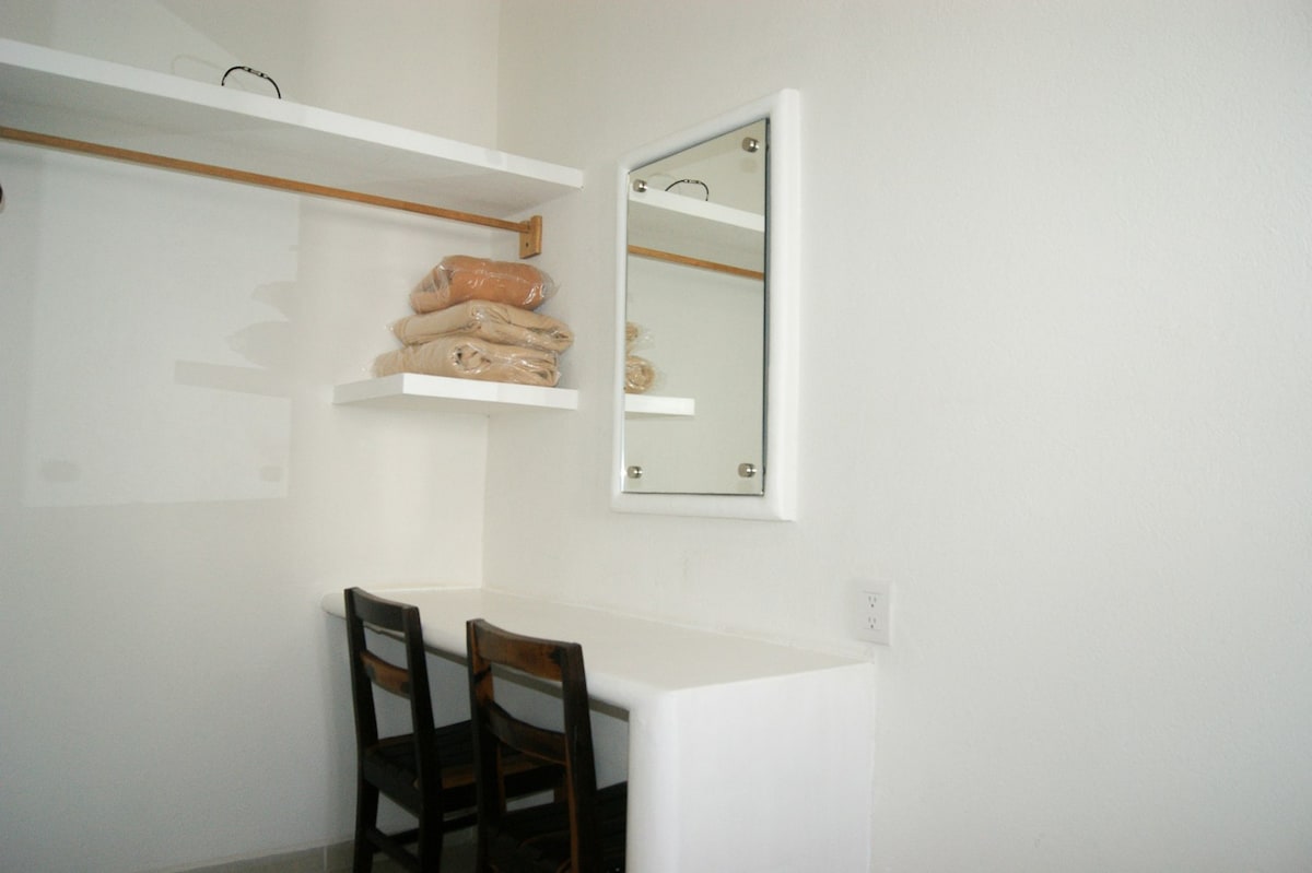 A compact area features a white wall with a mounted mirror and a simple table paired with two wooden chairs. Above the table, a shelf holds neatly stacked towels, providing an organized and functional space.