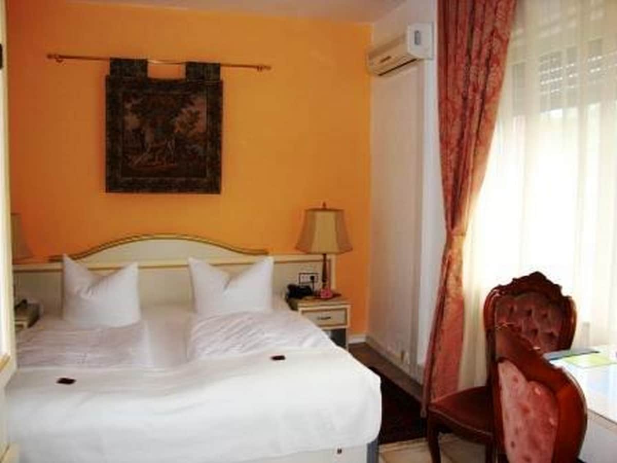 A cozy room features a spacious bed with white linens and decorative pillows. Two bedside lamps provide soft lighting. An elegant chair is placed near a desk, and large windows allow natural light to fill the space, complemented by light curtains.