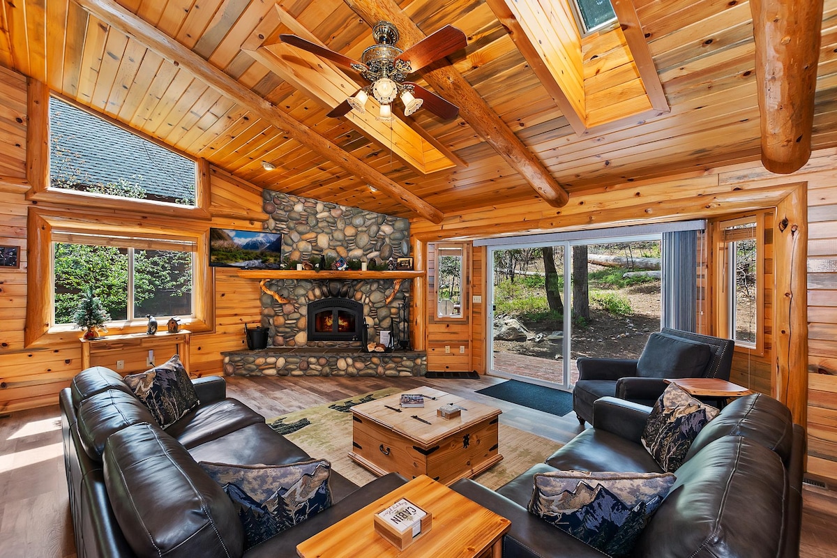 The living room features a stone fireplace surrounded by wood paneling, with large windows allowing natural light. Comfortable leather sofas and a wooden coffee table create an inviting seating area. The sliding glass doors open to a view of the outdoor space.