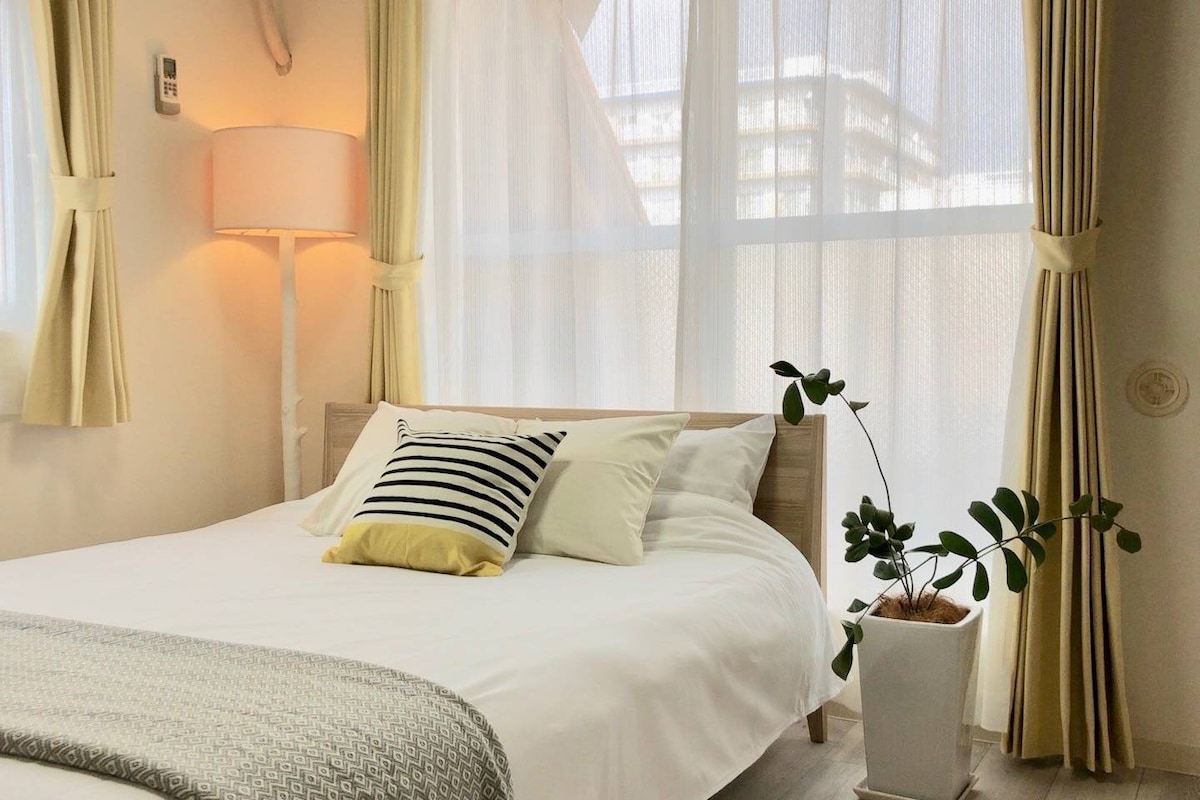 A well-lit sleeping area features a spacious bed adorned with a striped pillow and a decorative yellow accent. Sheer curtains allow natural light to filter in, while a small green plant in a white pot adds a touch of greenery to the room.