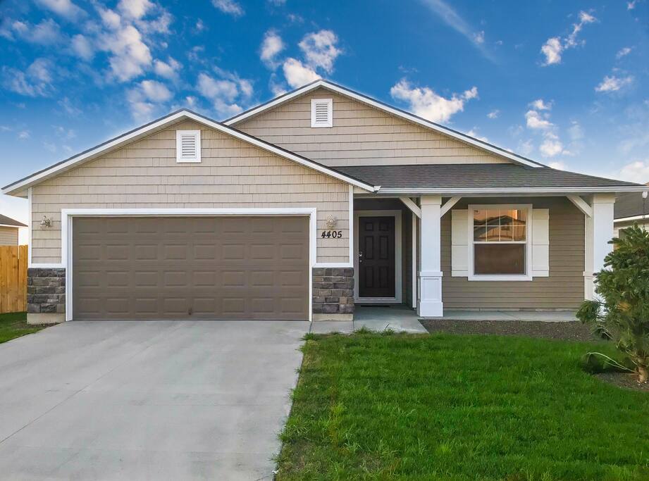 The exterior of the house displays a modern design with a combination of light-colored siding and stone accents. A spacious driveway leads to a two-car garage. The front porch is shaded by a roof, surrounded by a well-kept lawn and green landscaping.