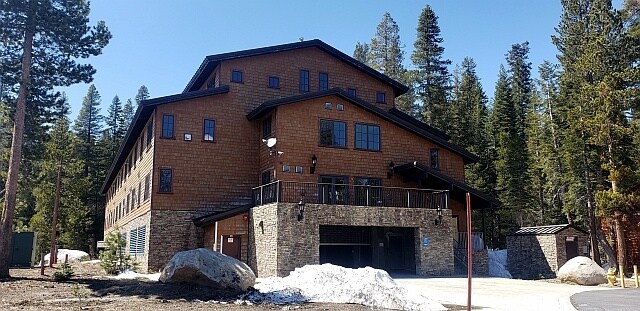 A three-story mountain condo building is surrounded by tall pine trees. The exterior features a blend of stone and wooden materials with large windows. A snow-covered area is visible in the foreground, highlighting the natural setting of Bear Valley Village.