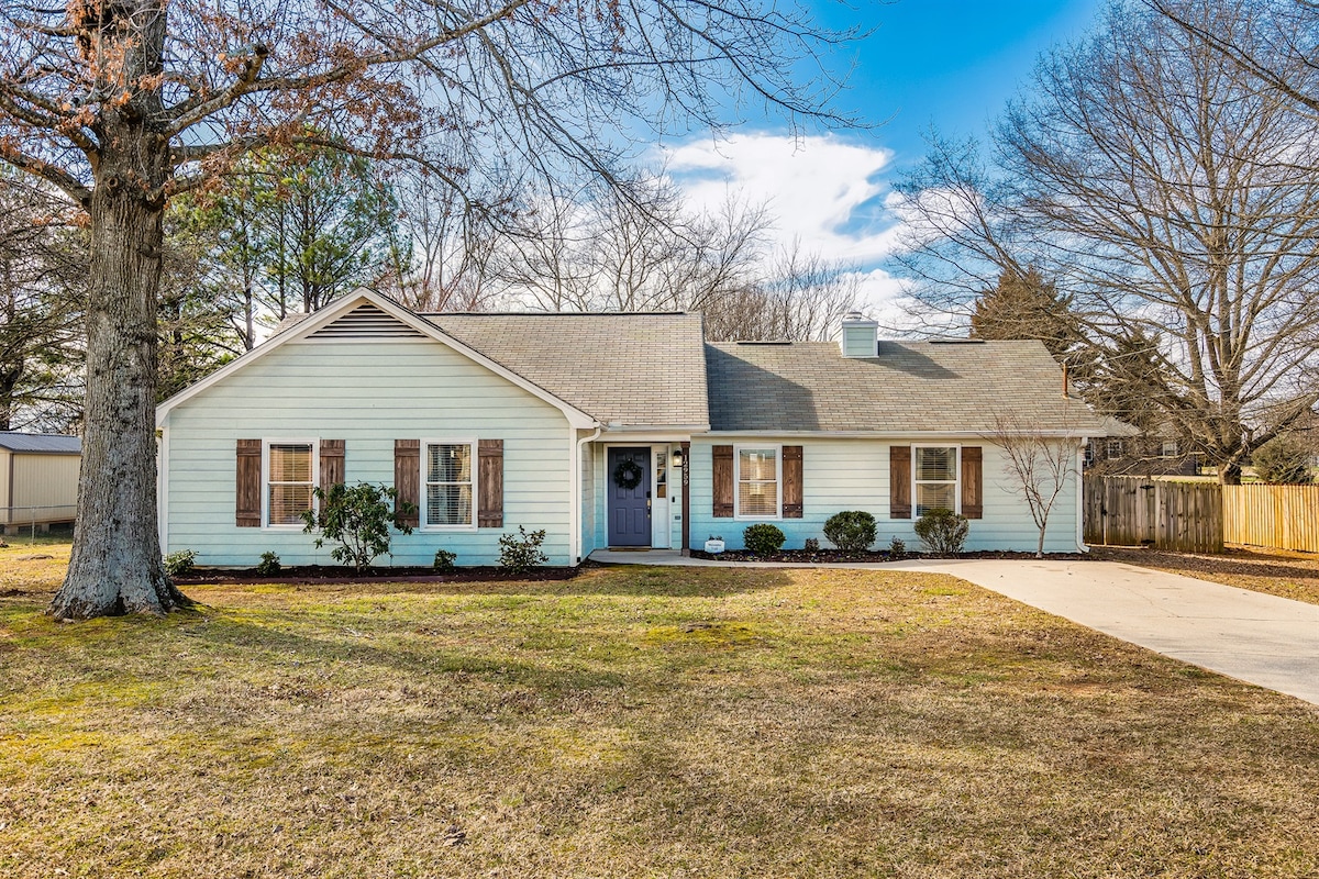 A single-story home with light blue siding and a welcoming entrance is surrounded by a well-maintained lawn. The front features decorative shutters and a circular driveway leading up to the front door, flanked by shrubs and small plants.