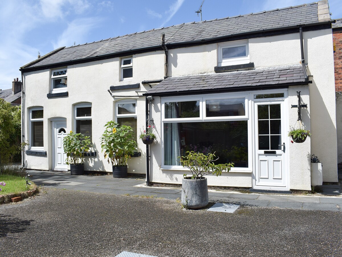 The exterior of a detached cottage is shown, featuring a combination of white and grey walls. A flat-roofed porch is visible at the front, with large windows allowing natural light into the living area. Potted plants are strategically placed for added greenery.