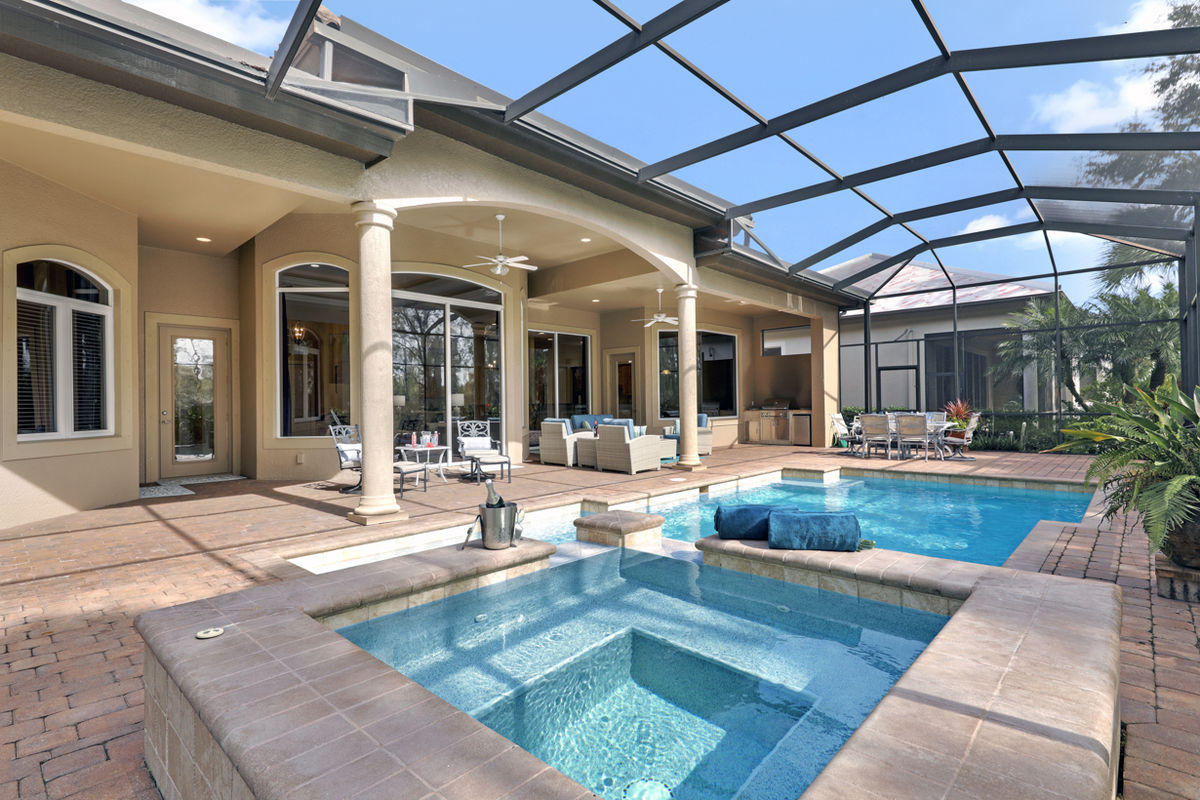 An outdoor area features a screened pool with a spa, complemented by a spacious patio. Comfortable seating is arranged nearby, surrounded by lush greenery. The architectural elements include tall ceilings and stone columns, offering an inviting and relaxing environment.