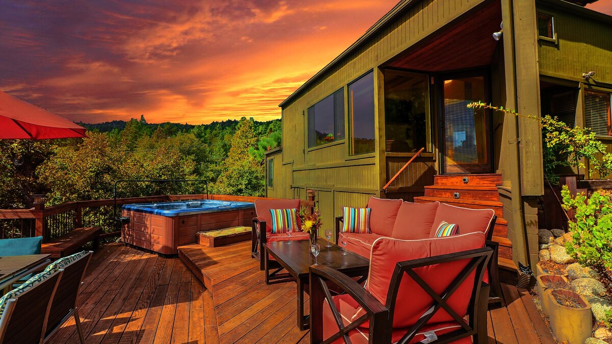 A spacious wooden deck is featured, accompanied by outdoor seating with comfortable cushions. A hot tub is situated close to the house, surrounded by greenery. A vibrant sunset paints the sky in warm hues, creating a serene atmosphere for relaxation.