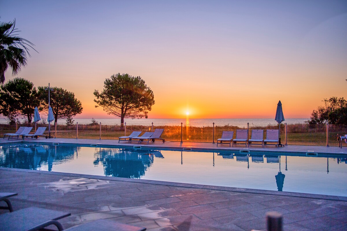 An outdoor heated pool reflects the vibrant colors of sunset, with lounge chairs positioned along the poolside. In the background, trees frame the scene, and the horizon reveals the sea at dusk.