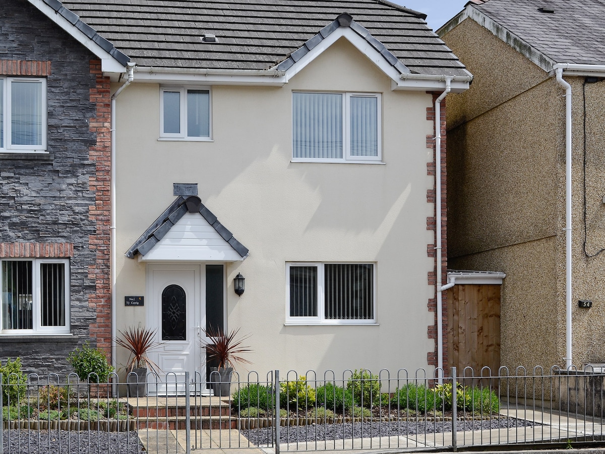 A semi-detached property is presented with a light exterior and decorative stone accents. A welcoming front door is framed by simple landscaping, featuring low shrubs and gravel. The property is complemented by a white picket fence and a neighboring house visible to the right.