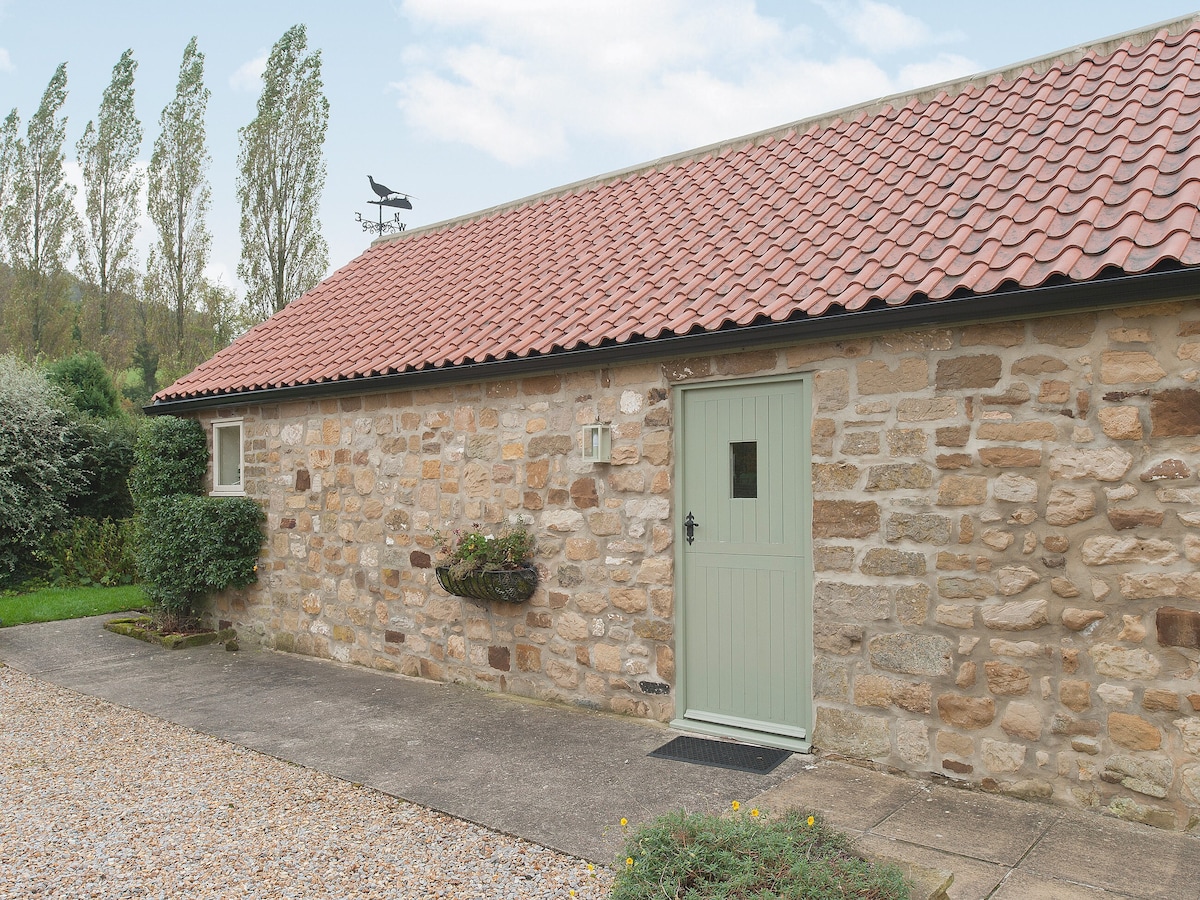 The exterior of a cottage is shown, featuring a stone façade and a traditional red-tiled roof. A light green door provides access, while a small plant holder is mounted beside the entrance. A gravel path leads to the front, surrounded by greenery.