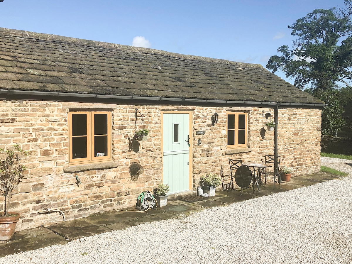 The exterior of The Old Cow Shed features natural stone walls and a subtle green door. Two windows allow light to brighten the façade, and flower pots adorn the entrance. A small seating area with a table and chairs is positioned beside the entrance, surrounded by gravel.