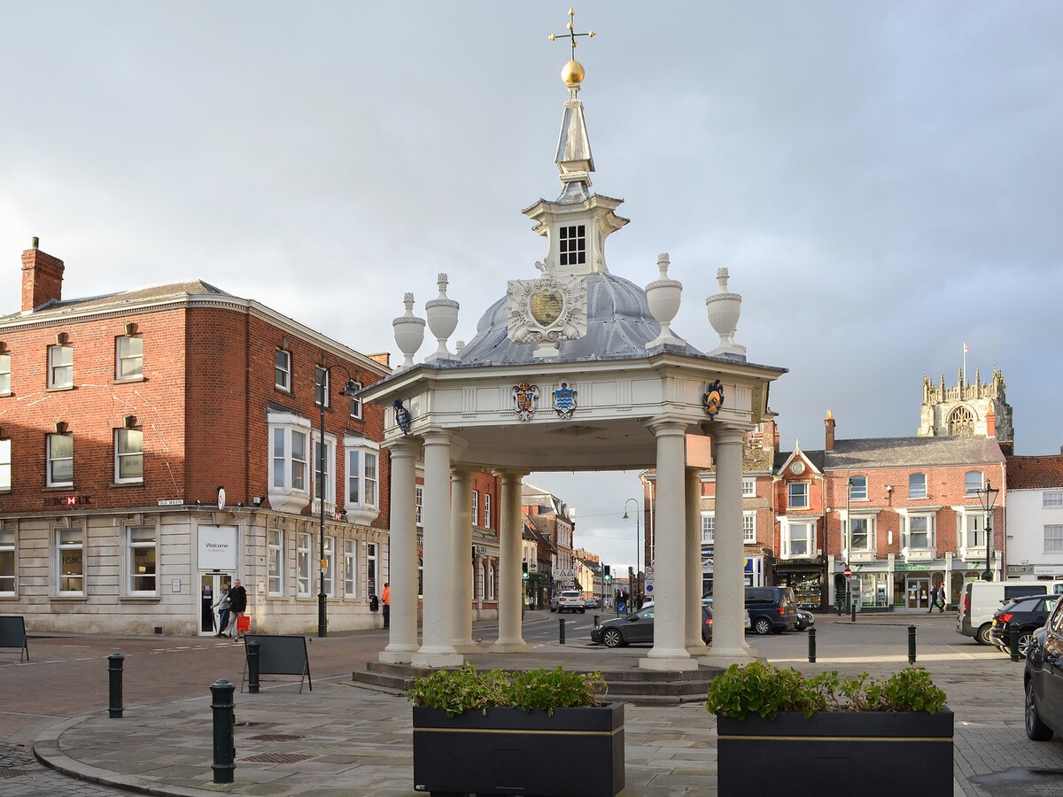 A historic structure with a decorative roof and columns stands prominently in a town square. Surrounding buildings reflect various architectural styles, showcasing brick facades and shopfronts. Planters with greenery enhance the area, while vehicles are parked along the streets, conveying a lively community atmosphere.