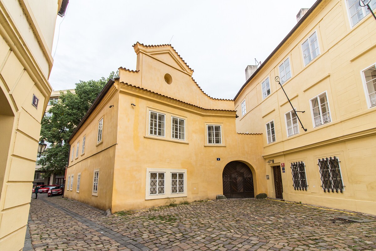 A charming apartment building with a light yellow facade is situated on a cobblestone street. The structure features a decorative roofline and several windows, reflecting a blend of historical architecture. Surrounding trees provide a touch of greenery in the tranquil courtyard area.