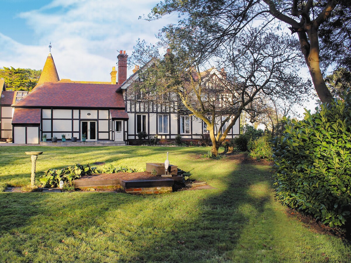 The exterior view of the property is showcased, displaying a two-story structure with distinct architectural features, including a turret and large windows. A well-maintained garden area includes a small water feature surrounded by greenery. The scene is illuminated by natural light under a clear sky.