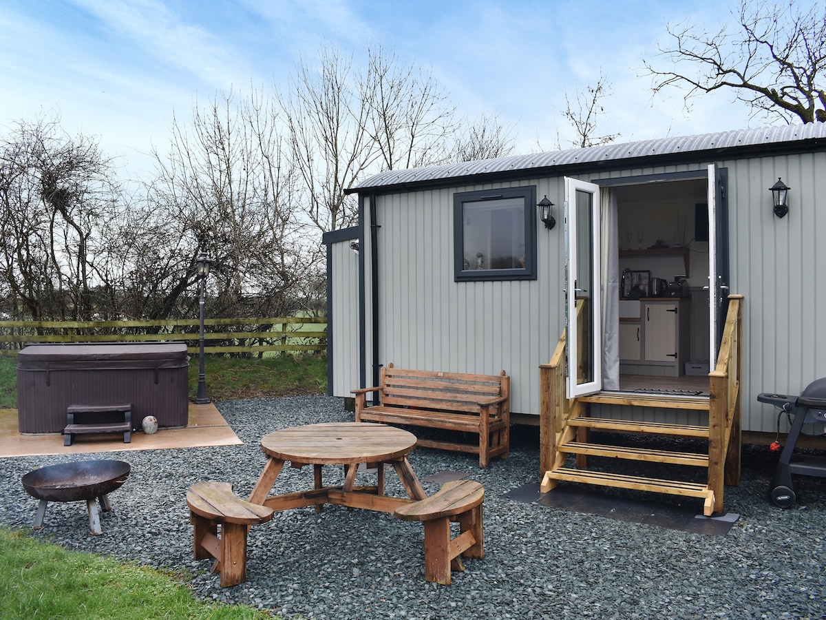 An outdoor area features a private hot tub and wooden seating arrangements, including a round picnic table and benches. Steps lead up to an open doorway, revealing the interior of the shepherd's hut. The surrounding landscape is open with sparse trees.