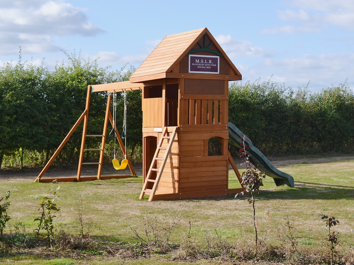 A wooden playhouse is situated in a grassy area, featuring a slide and swings. The structure includes a ladder leading to an elevated play space. Surrounding shrubbery adds a touch of greenery, and clear skies create an open and inviting atmosphere.