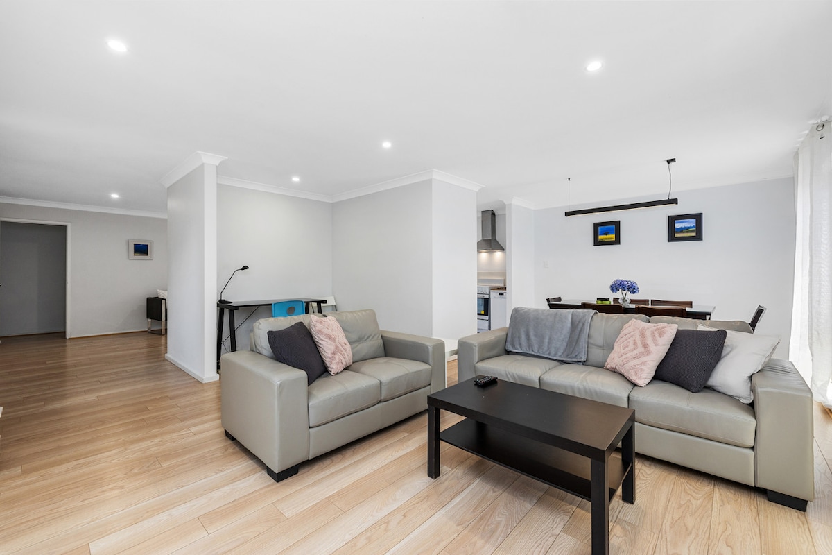 A modern open-plan living area features two comfortable sofas arranged around a dark coffee table. Natural light brightens the space, highlighting the light-coloured wooden flooring. A dining table for six is visible in the background, accompanied by contemporary wall art.