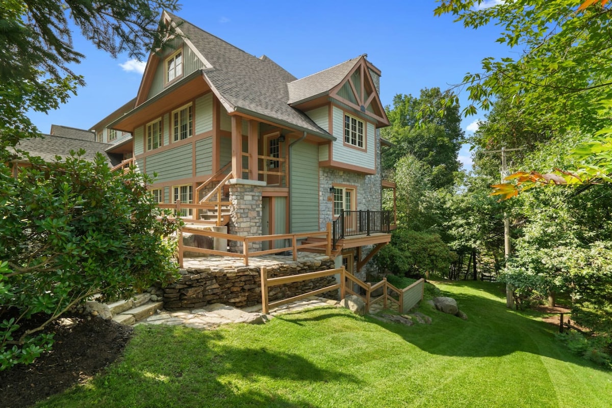 A charming exterior view of a multi-story house, featuring a stone and wood façade. A spacious wooden deck is visible, leading to a well-maintained grassy lawn surrounded by lush greenery, providing a peaceful outdoor setting.
