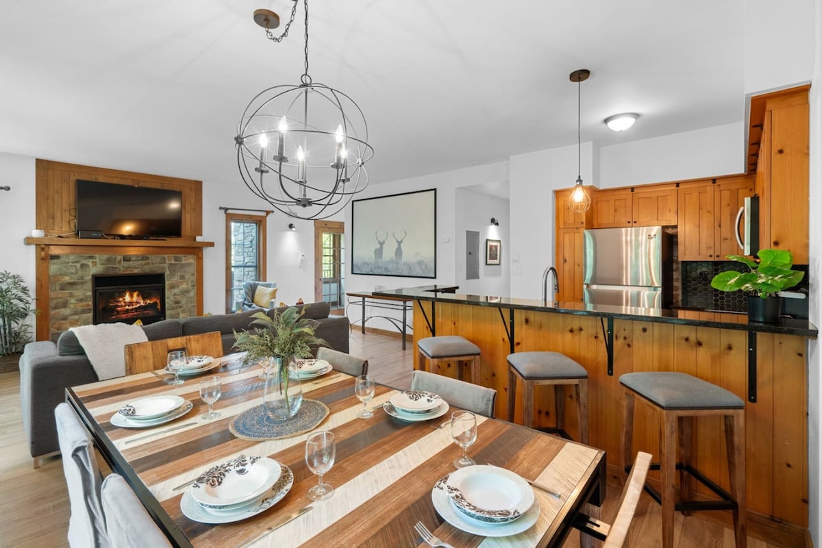 A spacious dining area features a striking wooden table set for six, with decorative plates and glasses arranged neatly. An inviting living space is visible in the background, showcasing a stone fireplace and modern furnishings. A large chandelier adds a warm glow to the atmosphere.