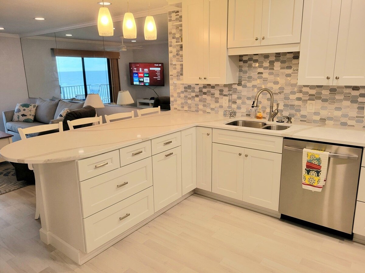 A modern kitchen area features sleek white cabinetry and a spacious countertop. Stainless steel appliances, including a dishwasher, complement the light-colored decor. A window allows natural light and provides a glimpse of the ocean view, while pendant lights illuminate the space.