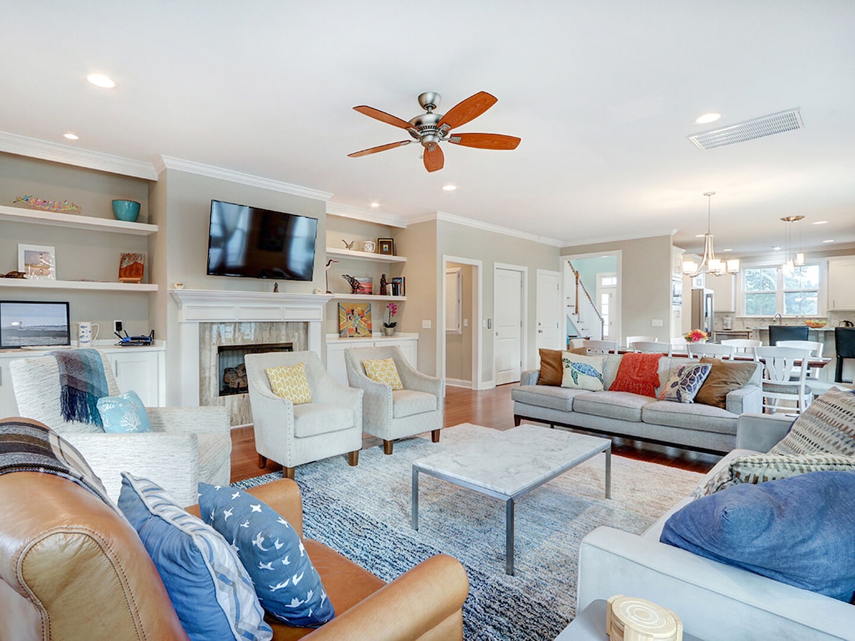 A spacious living area is showcased with plush seating arranged around a central coffee table. A flat-screen TV is mounted above a decorative fireplace. Light wood flooring enhances the open layout, with a dining area visible in the background, creating a welcoming space for gatherings.