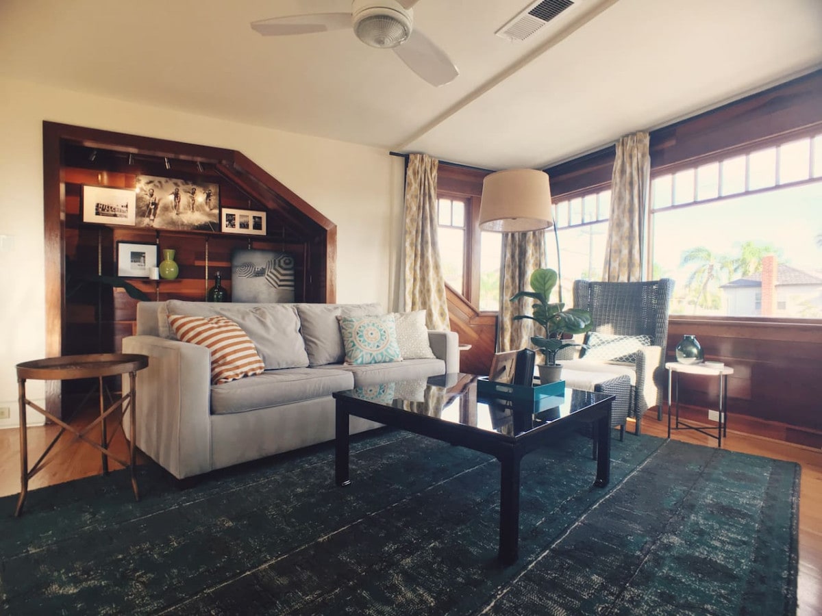 A light-filled living area features a comfortable gray sofa adorned with decorative cushions. A sleek black coffee table sits in front, complemented by a woven chair and side table. Large windows offer views of greenery, while the warm wood paneling adds character to the space.