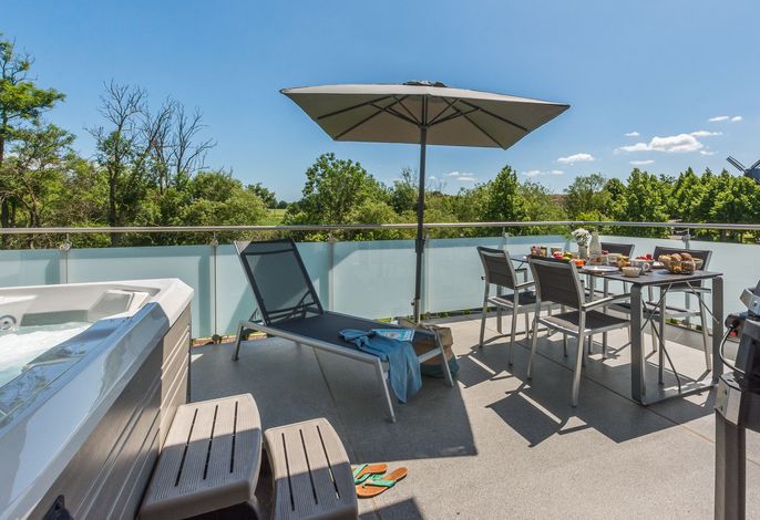 An outdoor space features a spacious terrace with a dining table set for several guests. A comfortable lounge chair and a large umbrella provide shade. The scene includes a hot tub and is surrounded by greenery against a clear blue sky.