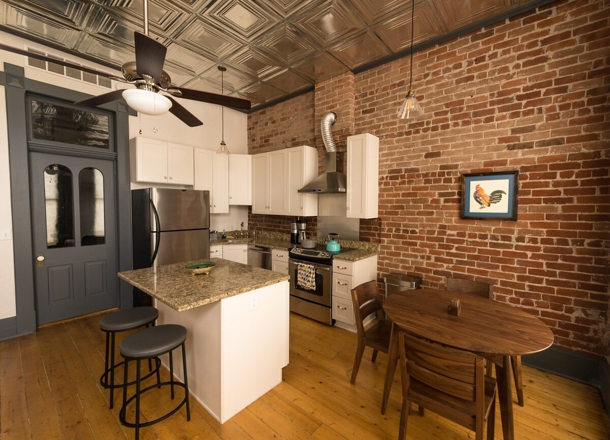 A spacious kitchen features modern appliances, including a stainless steel refrigerator and oven. A central island with bar stools is complemented by a wooden dining table. Exposed brick walls and unique tin ceiling tiles are highlighted, creating a blend of historic and contemporary design.