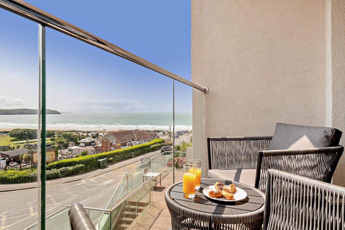 An outdoor balcony features a circular table with two black chairs, offering a clear view of the coastline and sea. A glass of orange juice sits on the table alongside a plate of pastries, with the horizon framed by a bright blue sky.