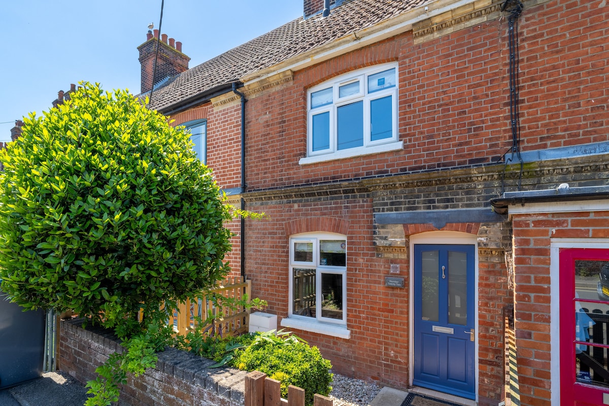 The exterior of a terraced cottage is presented, showcasing a red brick facade complemented by white window frames. A neatly trimmed hedge and small front garden create an inviting appearance. The entrance is marked by a blue front door, leading to a cozy interior.