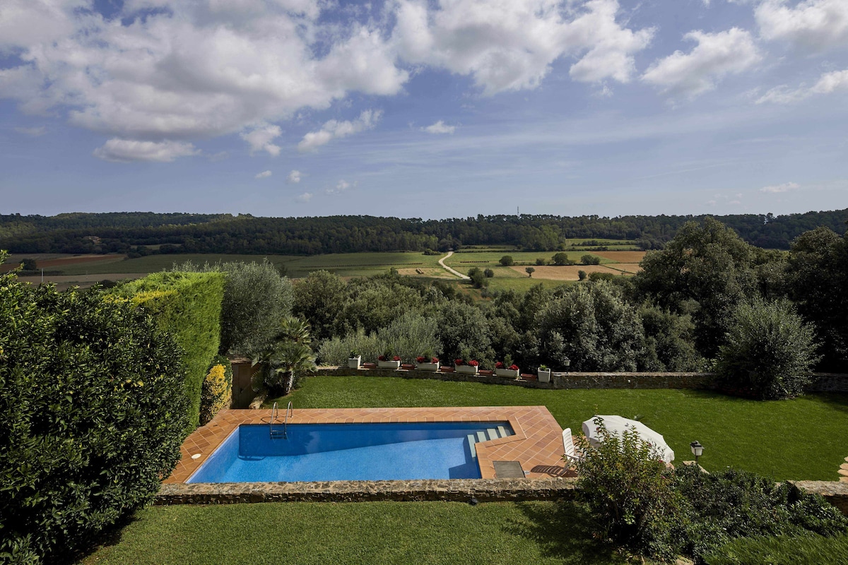 A serene outdoor space features a swimming pool surrounded by well-maintained gardens and trees. The landscape unfolds beyond with rolling hills and fields, under a partly cloudy sky. Lounge chairs and an umbrella provide shaded seating by the poolside.