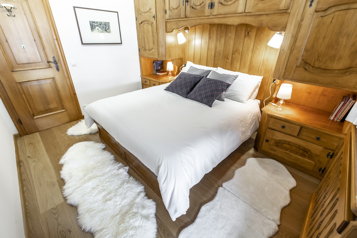 A warm bedroom features a spacious bed with crisp white linens, accented by dark plaid pillows. Wooden paneling forms a rustic backdrop, complemented by two bedside lamps. Soft area rugs are placed beside the bed, enhancing comfort and warmth.