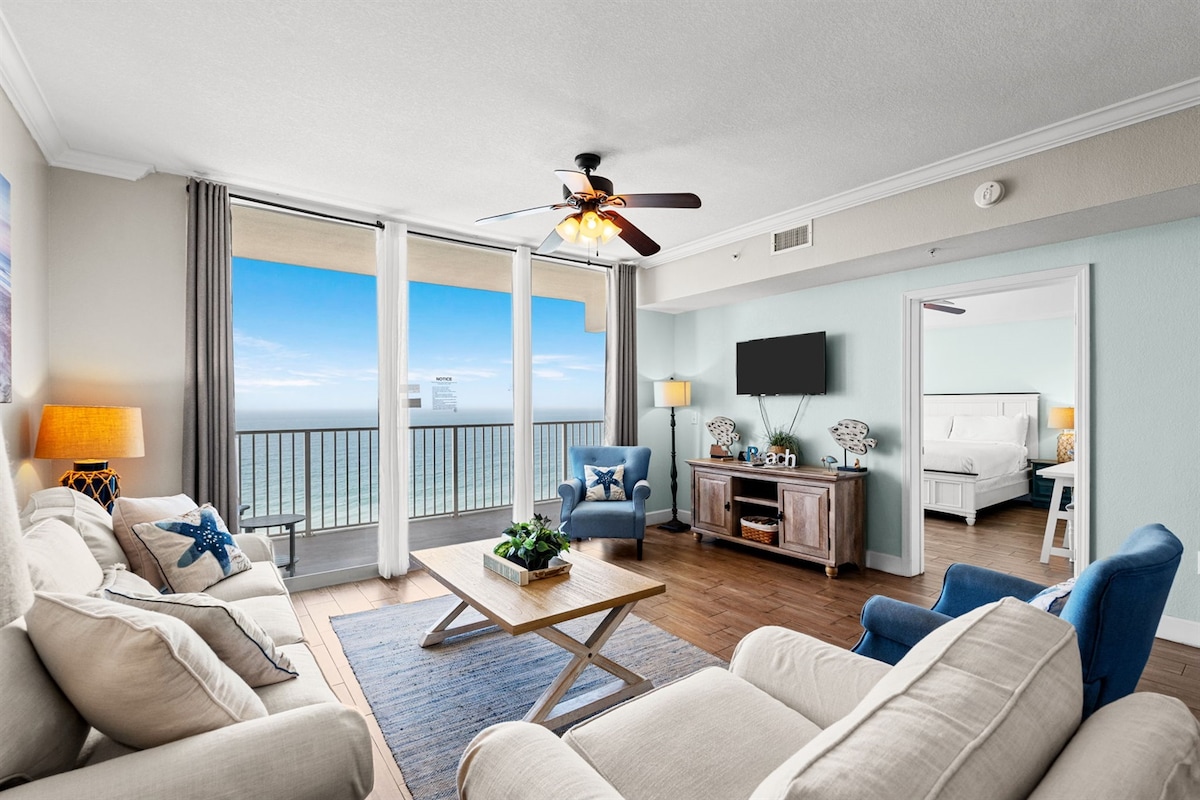 A bright and airy living area offers unobstructed Gulf views through expansive glass doors. Two comfortable sofas and a cozy armchair are arranged around a central wooden coffee table. A modern TV is mounted on the wall, with nautical decor adding a calming touch.