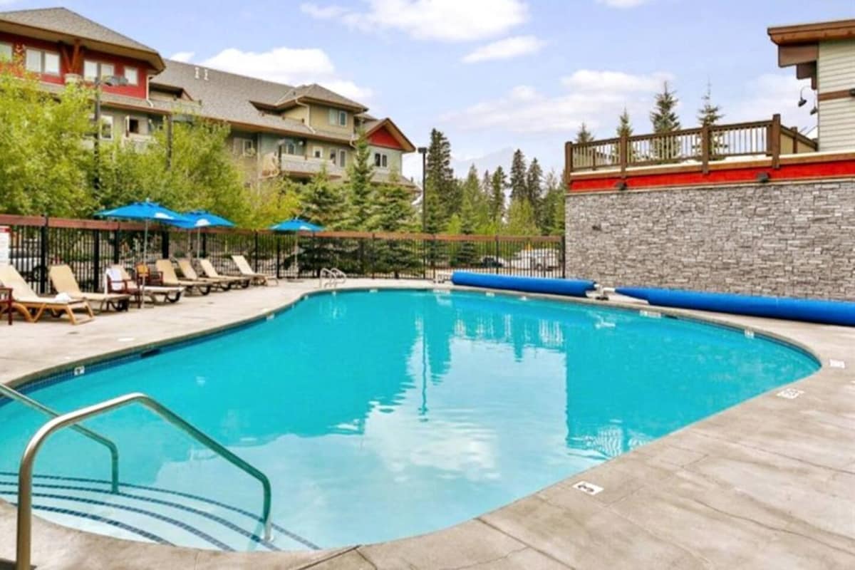 An outdoor heated swimming pool is set in a well-maintained area, surrounded by lounge chairs and umbrellas. A smooth concrete deck leads to the clear water, with trees and buildings providing a scenic backdrop.