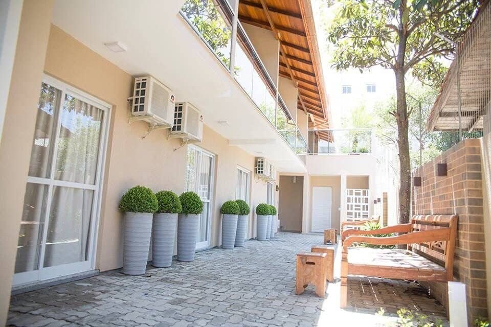 The exterior of the building features a paved area lined with tall planters containing greenery. Air conditioning units are mounted on the walls. A wooden bench is situated to the side, providing a shaded spot to relax.