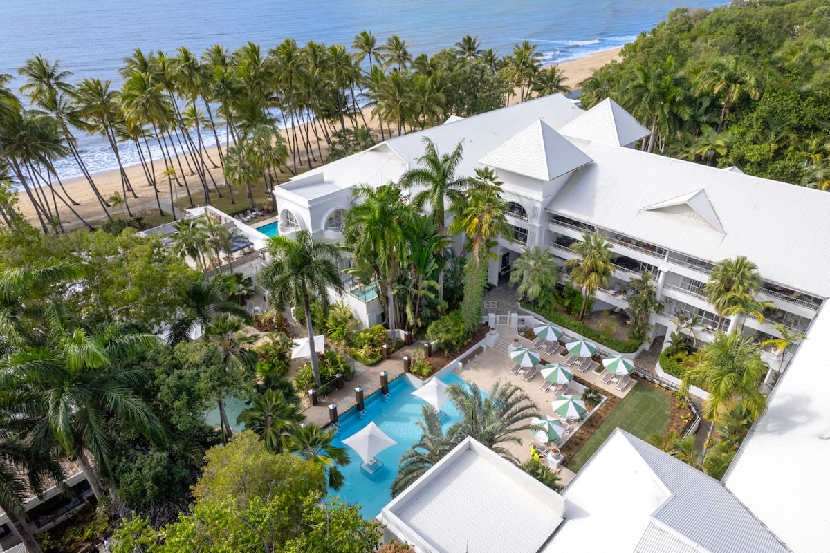 Aerial view showcases the resort's beautifully landscaped grounds, featuring lush palm trees, multiple swimming pools, and sun loungers. The coastline stretches in the background, highlighting the beachfront location, with golden sands and gentle waves visible.