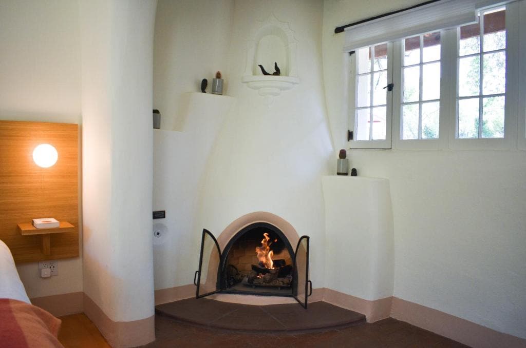 A serene corner of the room showcases a charming kiva fireplace, where a soft flame flickers. The white walls, complemented by rustic accents, create a warm ambiance. A series of small decorative items are arranged on a shelf, while natural light filters in through multiple windows.