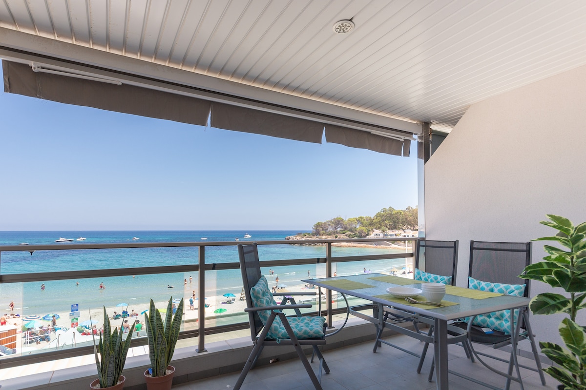 A private terrace offers stunning views of the Mediterranean Sea, with light blue waters and beachgoers visible below. A dining table is set with place settings, accompanied by several chairs. Potted plants line the edge, contributing to the outdoor ambiance.