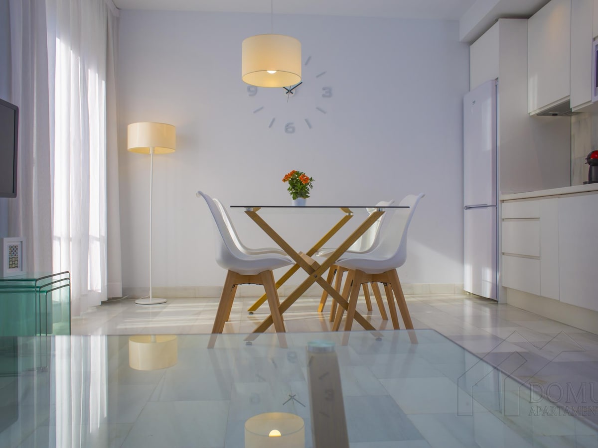 A modern dining area features a glass table surrounded by four white chairs, positioned under a contemporary light fixture. A small plant is centered on the table, and a minimalist clock adorns the wall. Natural light filters through sheer curtains, enhancing the space.