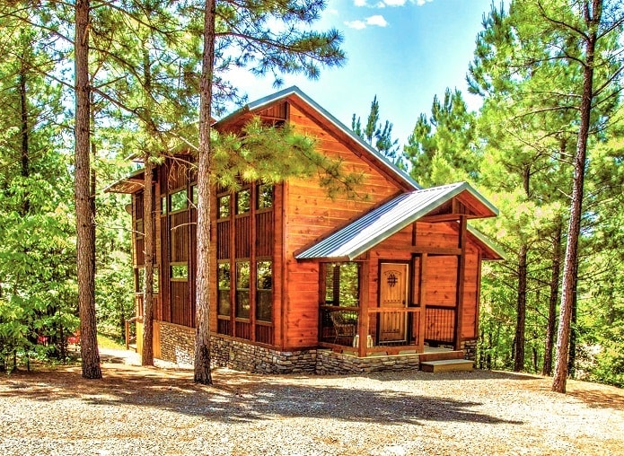 The exterior of the cabin showcases a contemporary log cabin design. Large windows are framed by wooden accents, allowing natural light to fill the interior. Surrounded by tall pine trees, a welcoming porch with stone detailing enhances the inviting nature of the property.